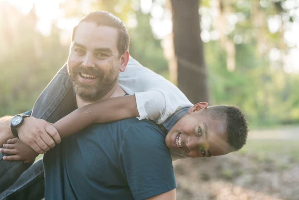 Smiling white man playfully slinging a smiling black boy over his shoulder, with sunlit forest in background