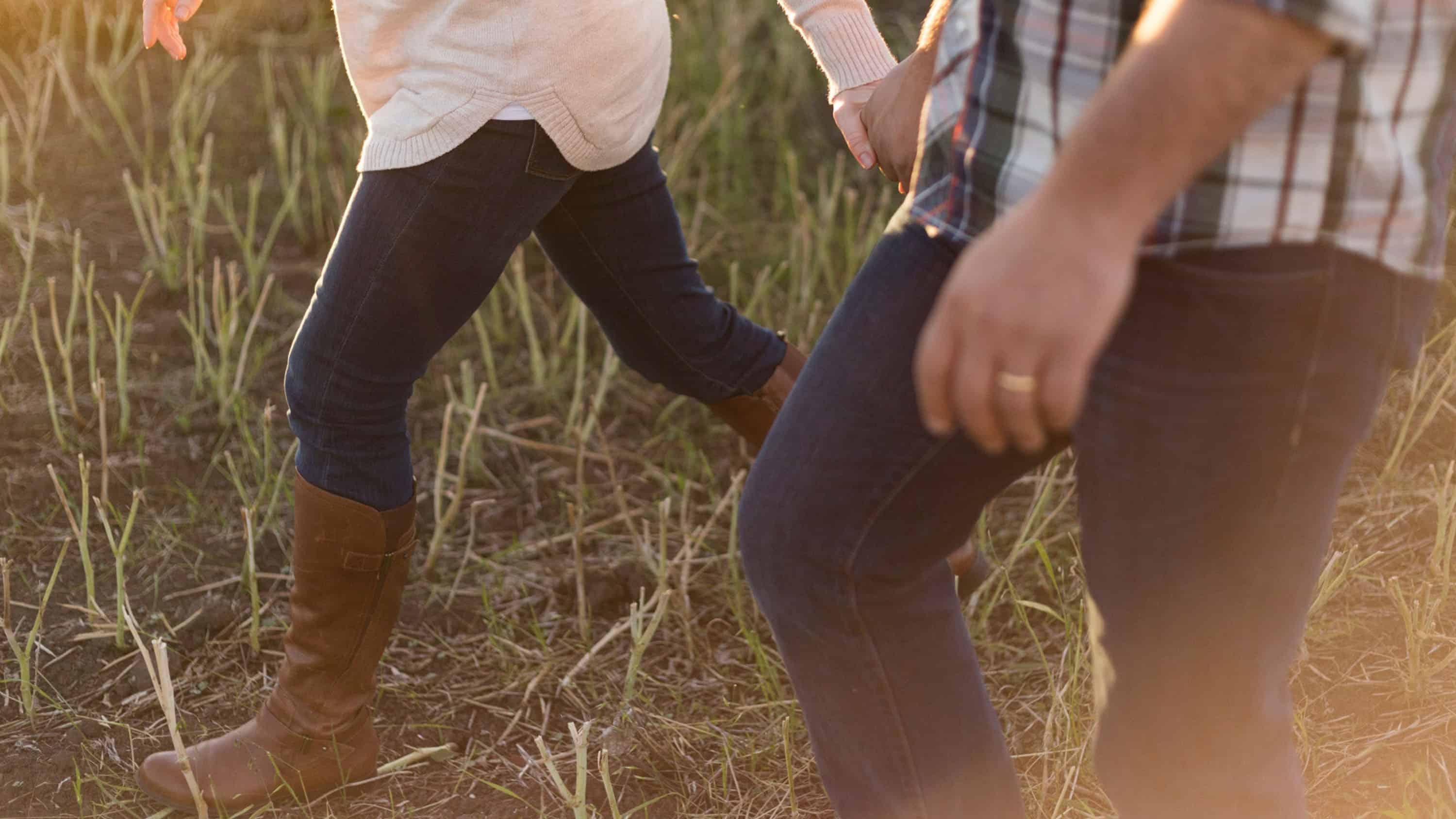 Close up of the legs of a married couple walking through a field. They’re both wearing jeans and holding hands.
