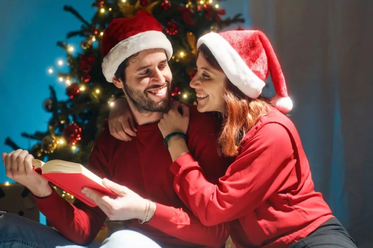A husband and wife wearing Santa hats and red sweaters share a warm moment, engaging in advent devotions for couples by the light of a Christmas tree.