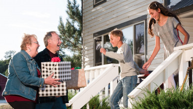 Happy grandparents arriving to their grandkids' home with presents in hand while two happy grandkids run out to meet them