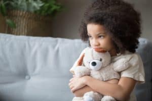 Sad-looking, young African-American girl sitting on a couch, hugging her teddy bear