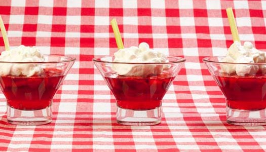 Three small glasses filled with a red jello-like dessert sitting in a row on a red and white checkered tablecloth