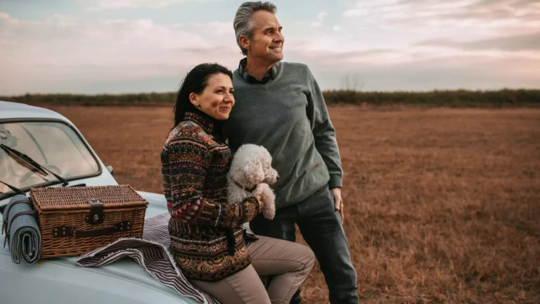 A happy middle-aged couple, now empty nesters, enjoy a peaceful outdoor moment with their small dog, leaning against a vintage car with a picnic setup.