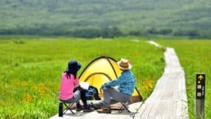 Husband and wife seated at a small campsite next to a wooden path in an open field. They’re staring at the hills beyond.