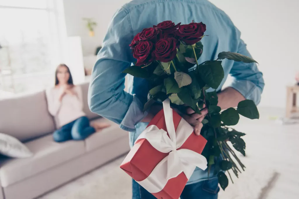 A photo of a man hiding a bouquet of roses and a gift box behind his back as a surprise for Valentine's Day.