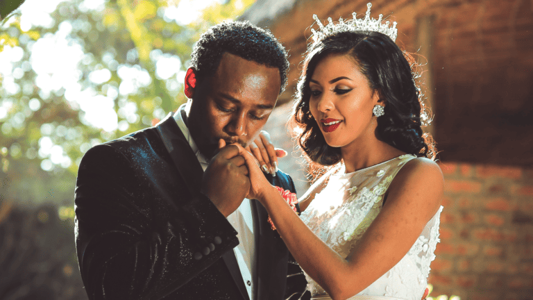 Young, black newlywed couple standing outside. He's holding her hand and kissing it.