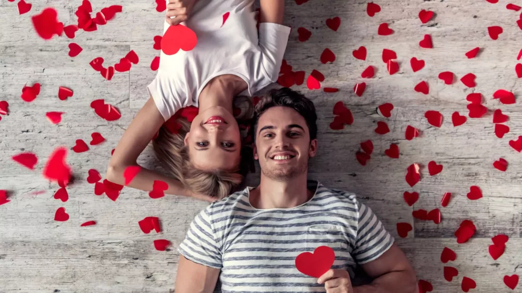 Photo of man and woman laying on the ground, each holding a paper heart over their chest, while paper hearts are sprinkled on them from above. They are happy because they have the secrets of healthy sex in their marriage.