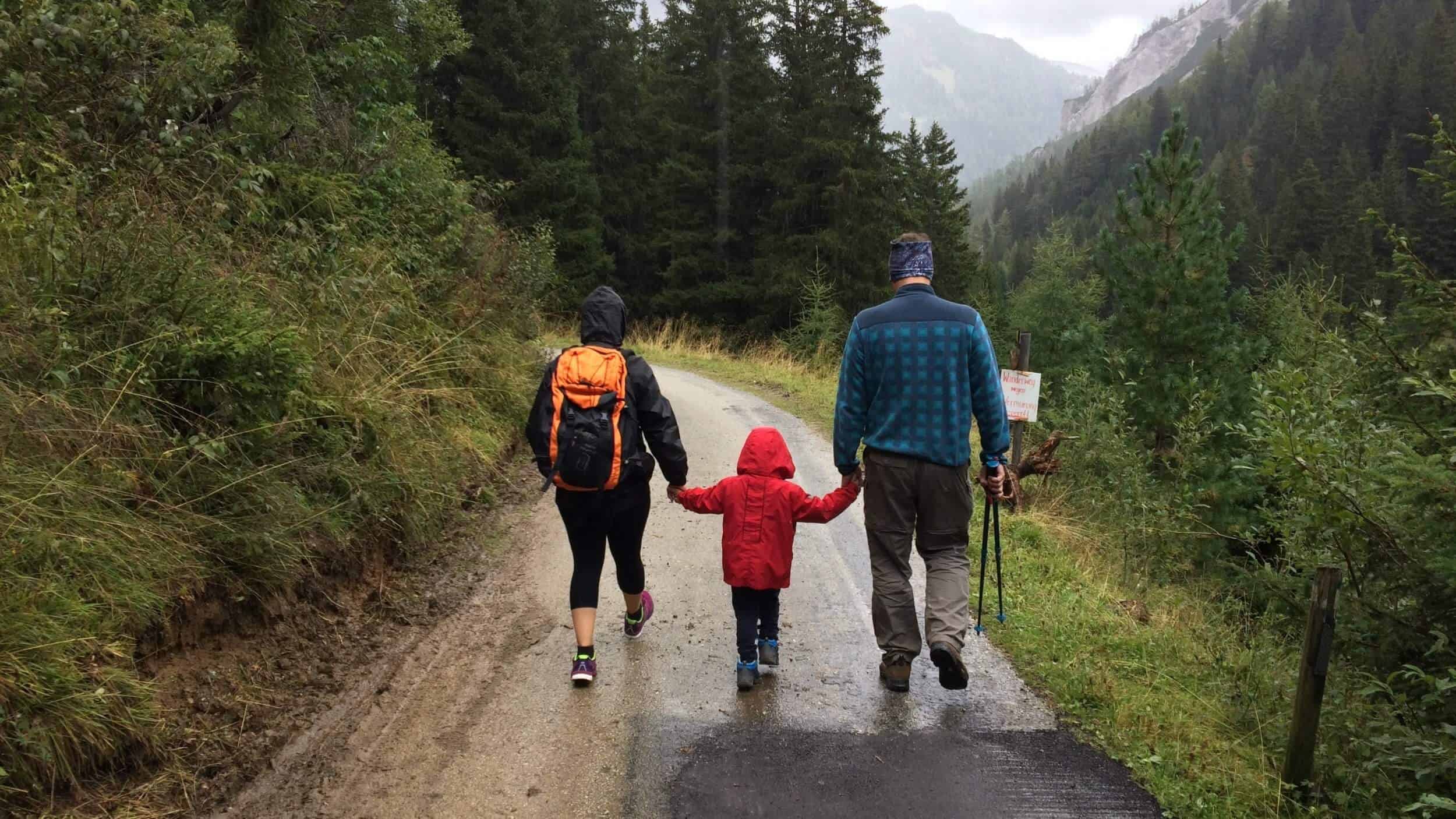 Shown from behind – a couple walking and holding hands with their young child on a mountain path on a rainy day