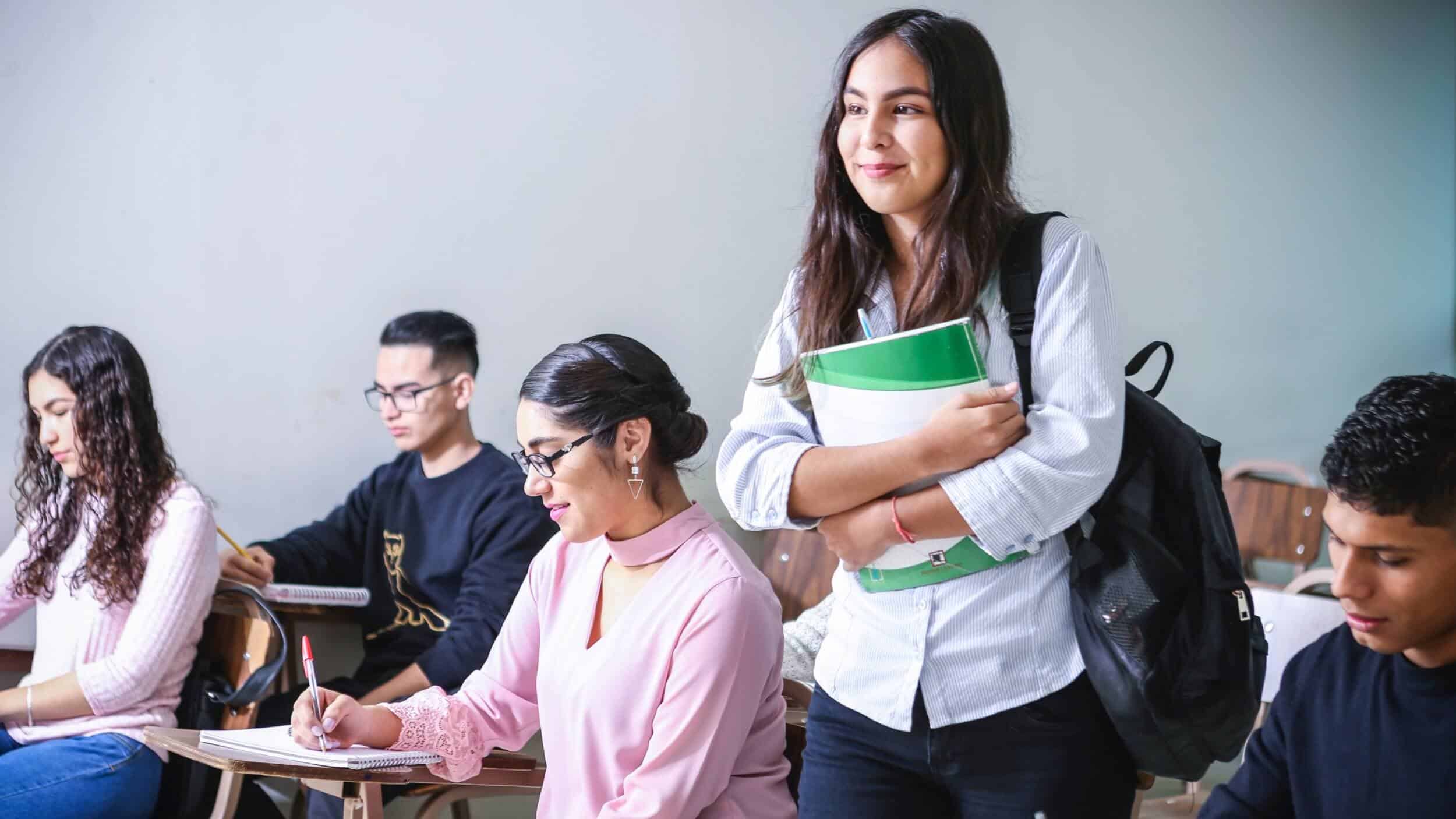 Confident, smiling high school girl standing in her classroom, surrounded by classmates working at their desks