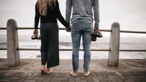 Shown from behind and the waist down, a barefoot husband and wife holding hands, looking out at the ocean