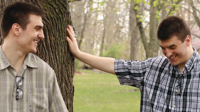 Two young men stand together by a tree laughing