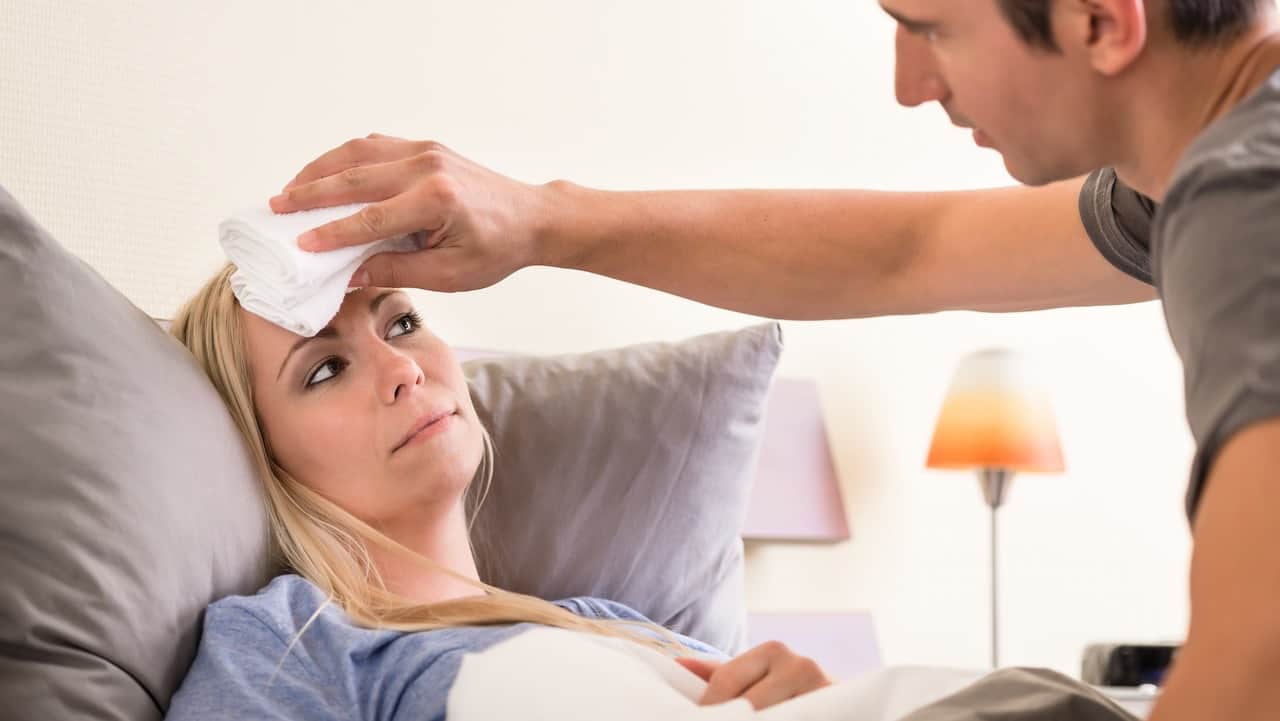 Ill wife lying in bed as husband applies a folded cloth to her forehead