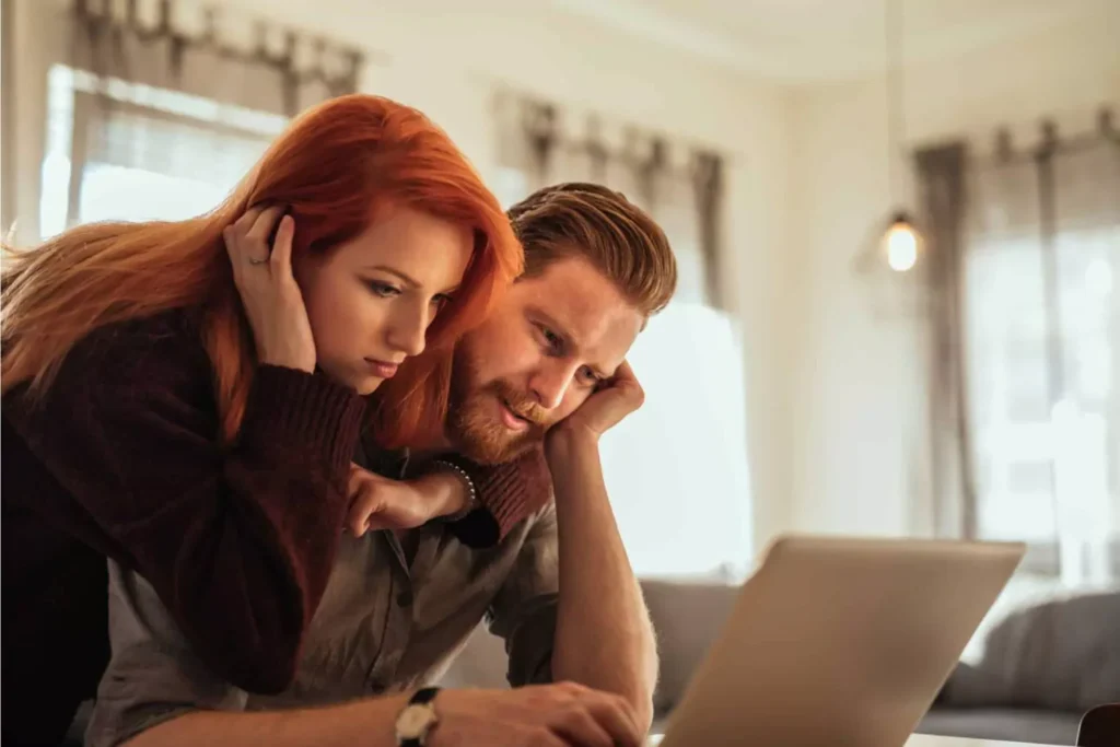 A couple sits closely together, focused on a laptop screen, discussing strategies for conquering the spending problem and managing their finances.