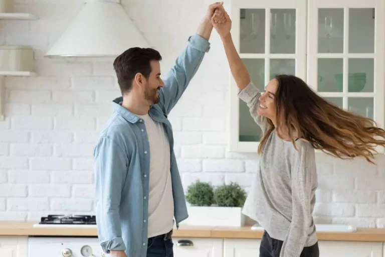 A loving husband twirls his smiling wife in the kitchen, symbolizing how to be a good husband by creating joyful and meaningful moments together.