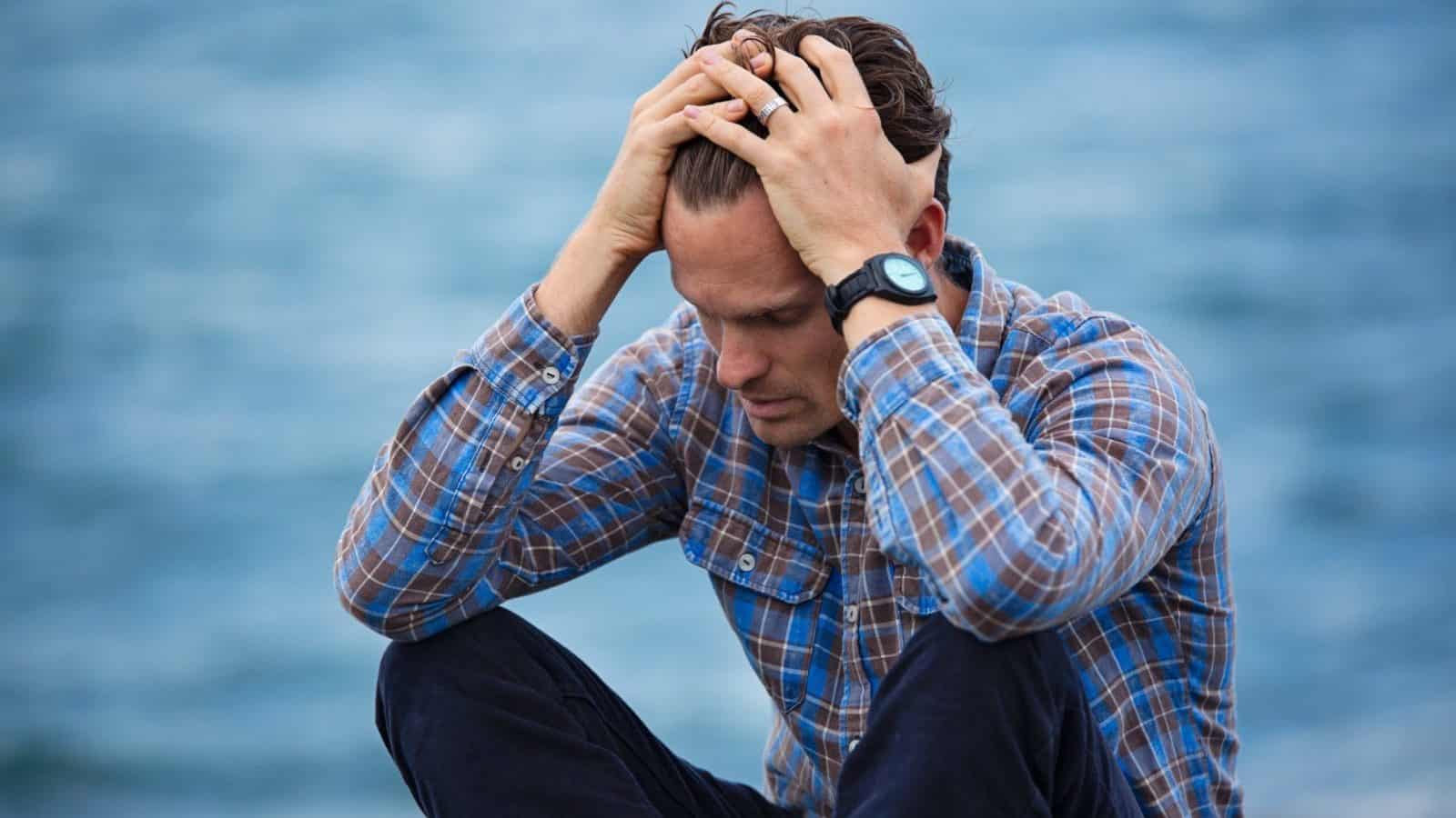 Upset looking husband sitting cross-legged in front of a body of water, with his head resting in his hands