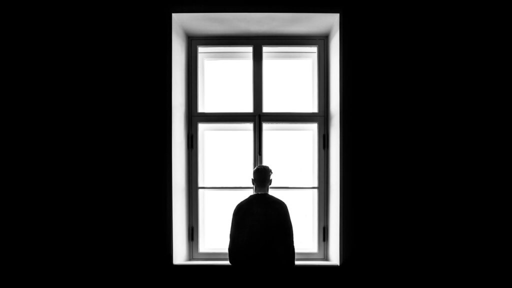 Black and white photo of man standing in the dark, looking out a brightly-lit window