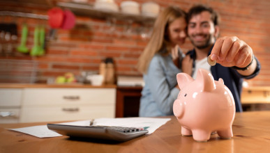 Ceramic piggy bank on a kitchen table in the foreground, with young couple adding a coin from the background