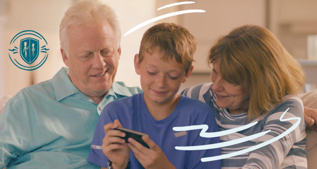 Boy flanked by his grandparents as they sit on a couch. They're all looking at a mobile device he's holding.