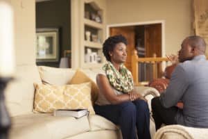 Upset middle age black couple sitting in their living room. He's talking to her, she's looking past him, avoiding his gaze