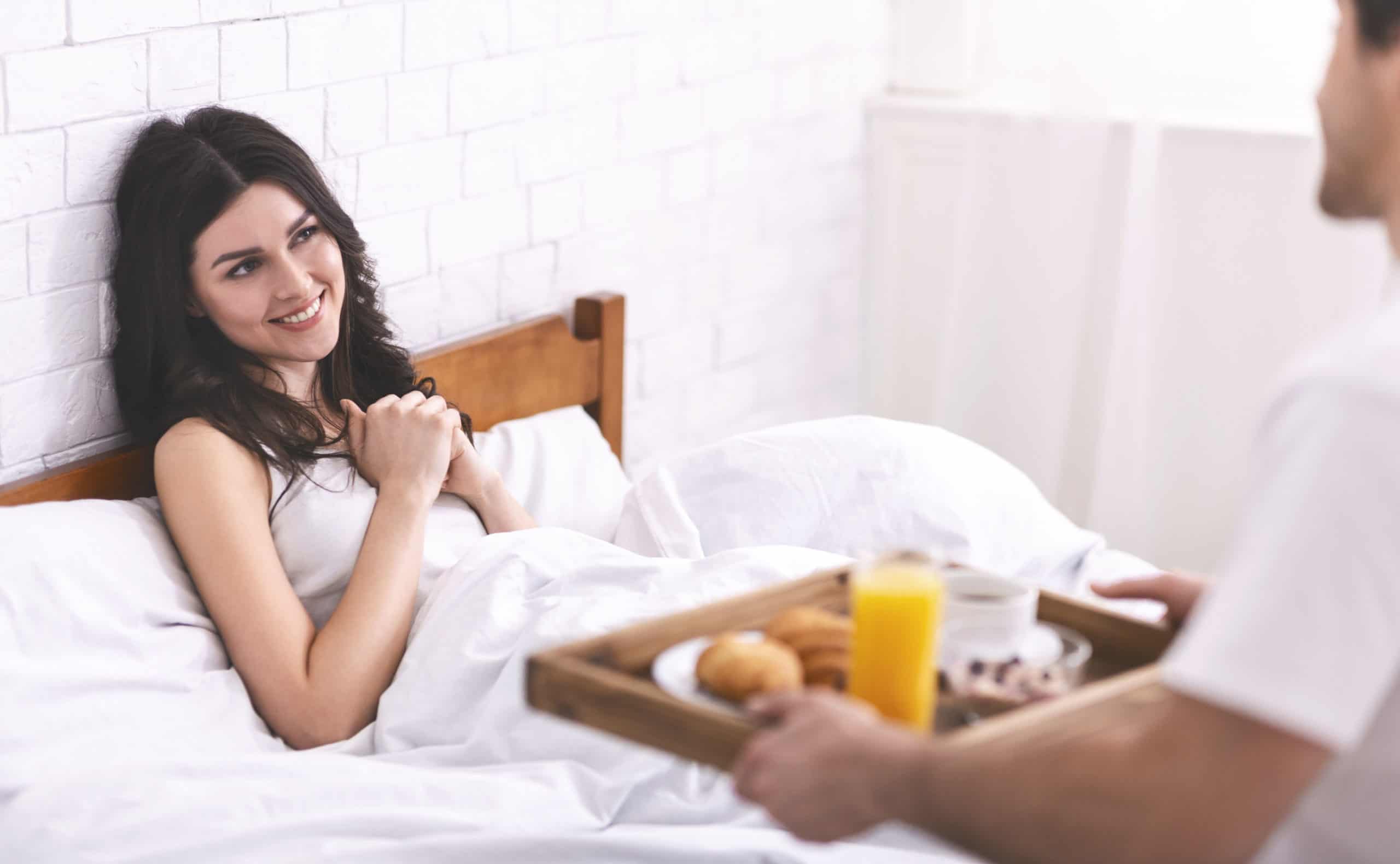 Smiling woman sitting up in bed as husband brings her breakfast on a tray