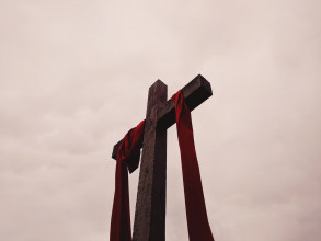 Upward-looking perspective of Christ’s cross with a red sash hanging on it and a cloudy sky as the background
