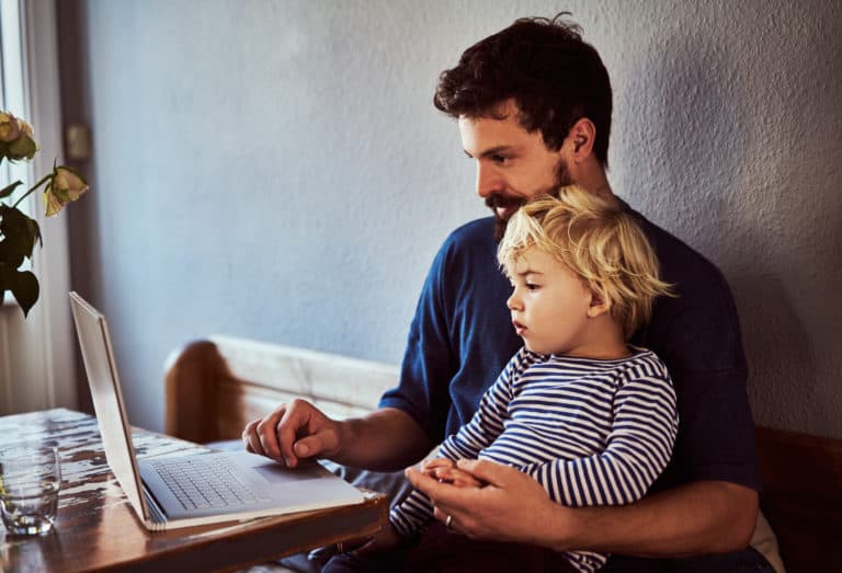 Young father sitting at a table looking at a laptop while holding his young son on his lap