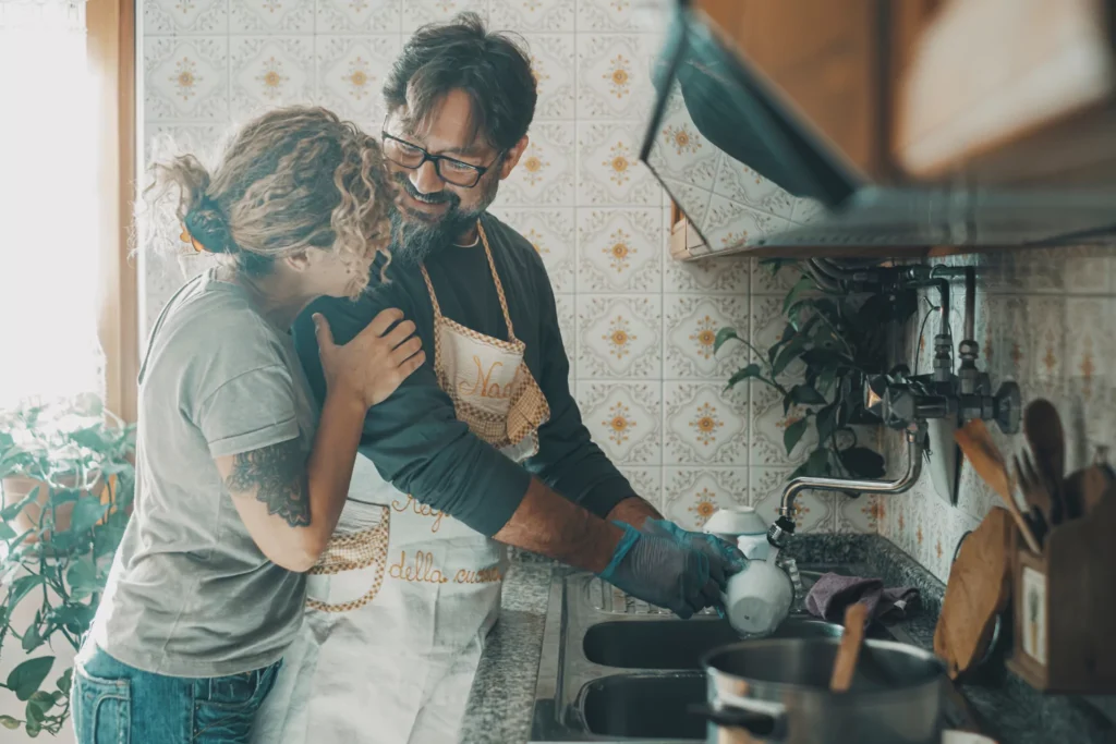 Man washing dishes at a kitchen sink while woman hugs him from behind, both smiling and enjoying a warm moment together.