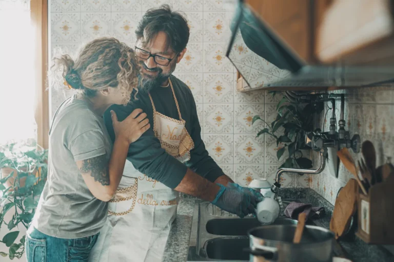 Man washing dishes at a kitchen sink while woman hugs him from behind, both smiling and enjoying a warm moment together.