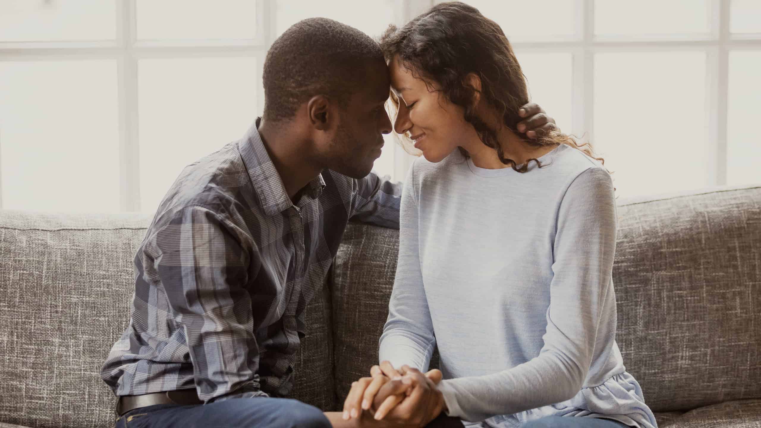 Young, affectionate African-American couple sitting on a couch holding hands, leaning head to head