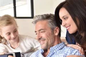 A happy couple sits closely together with their young daughter, sharing a warm moment while looking at a small box. The wife, with a gentle smile, leans on her husband’s shoulder, while he grins as he interacts with their child. Their expressions and body language convey affection, security, and mutual understanding, symbolizing the importance of building trust with your husband in a loving relationship.