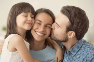 A joyful family moment where a husband kisses his smiling wife on the cheek while their young daughter hugs her, symbolizing the importance of honoring your wife by recognizing her value.