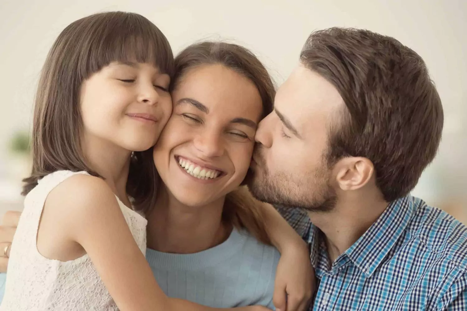 A joyful family moment where a husband kisses his smiling wife on the cheek while their young daughter hugs her, symbolizing the importance of honoring your wife by recognizing her value.