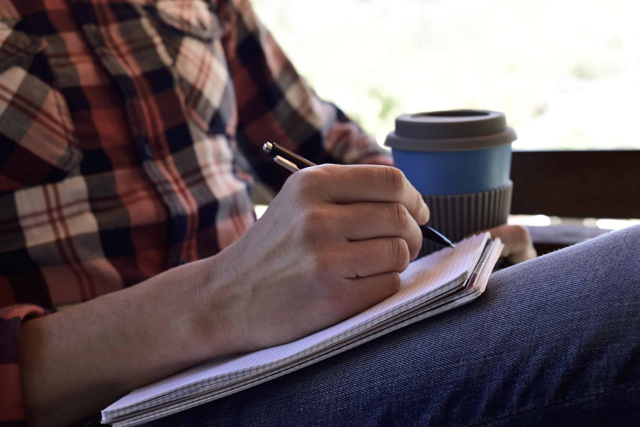 Close up of a man's hand writing in a notebook; he's casually dressed and holding a cup of coffee in the other hand