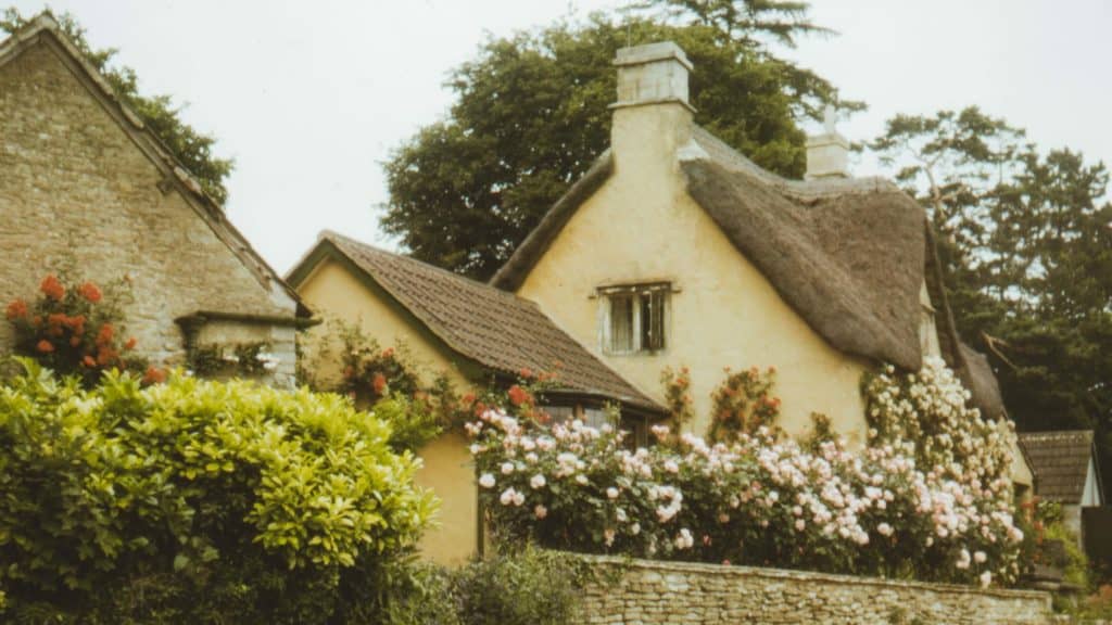 Slightly faded photo of an old-looking stone cottage surrounded by flowers, greenery, and trees