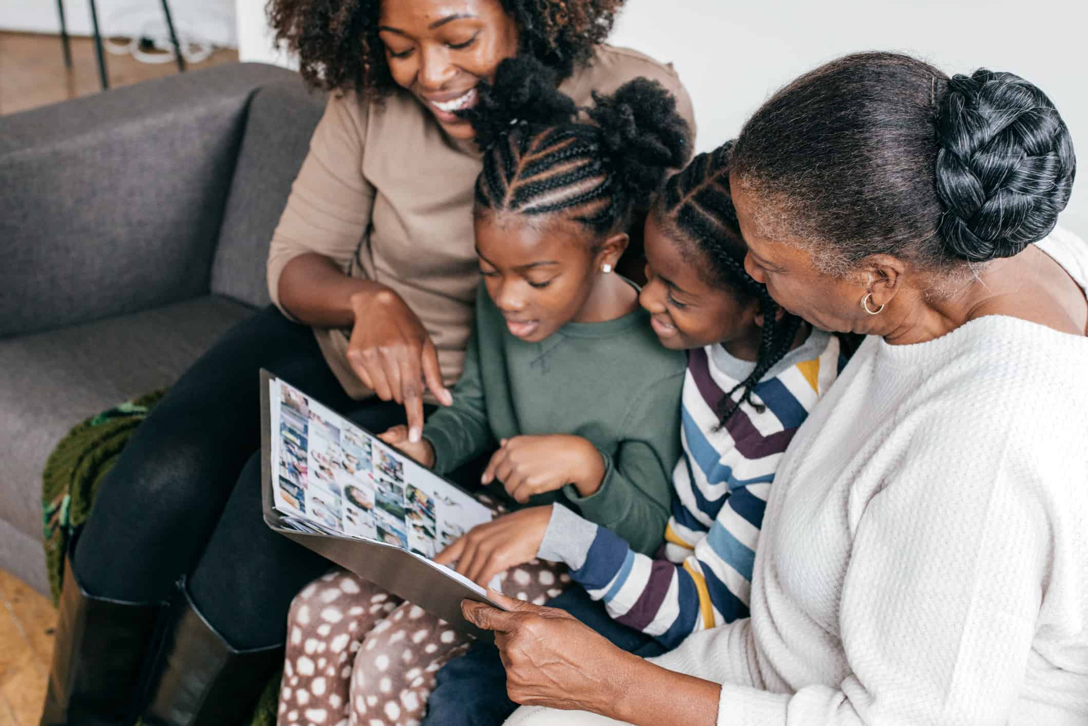 Black family of mom, two young daughters, and grandmother all sitting on a couch enjoying looking at a photo album