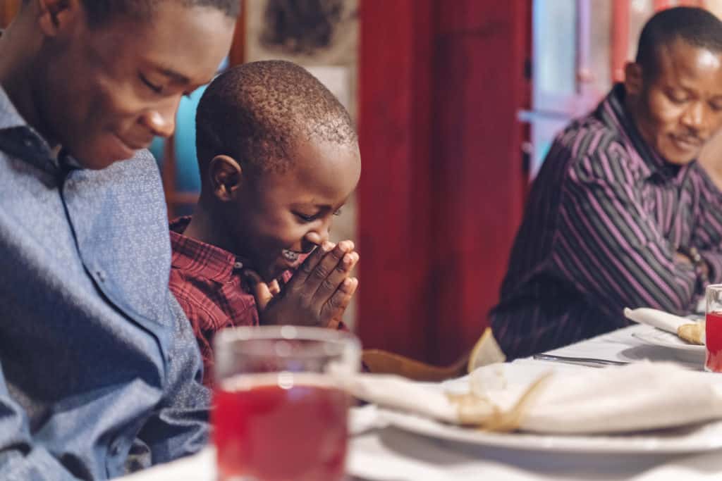 Black family at a dinner table; close up of dad and young son smiling with their heads down and eyes closed for prayer