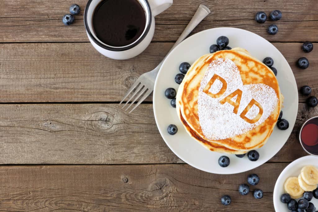Stack of pancakes next to cup of coffee. Top is sprinkled with powdered sugar in shape of a heart with the word 'dad' in it