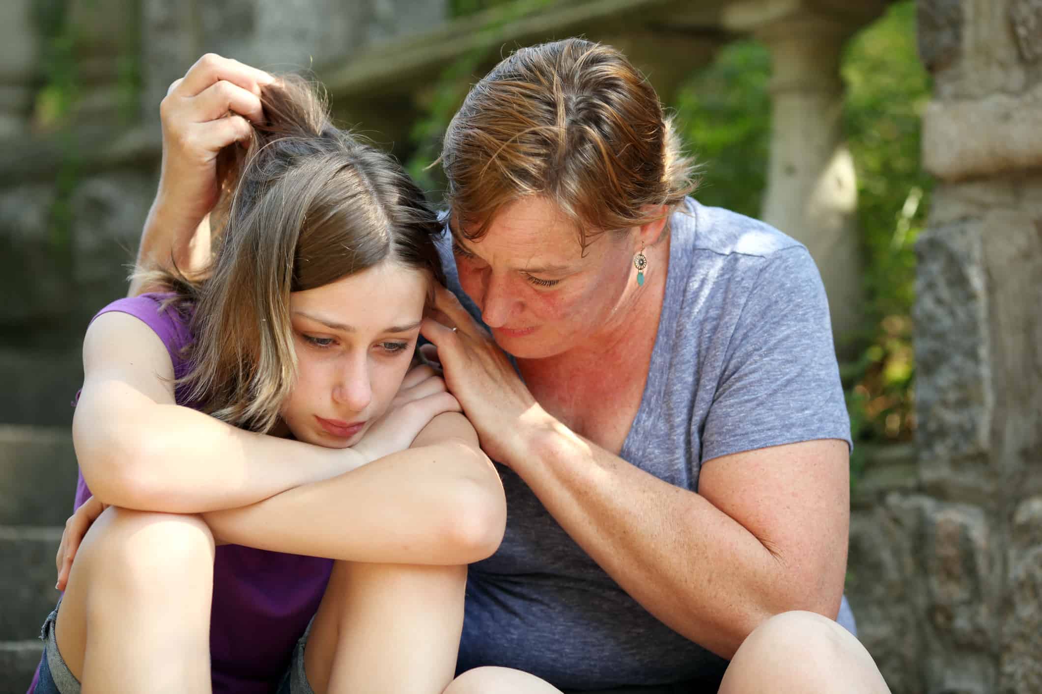 Mom affectionately comforting her sad tween daughter as they sit together on some steps outside