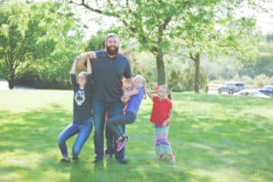 Smiling dad with his son and three daughters in a park; one daughter is hanging off each of his arms