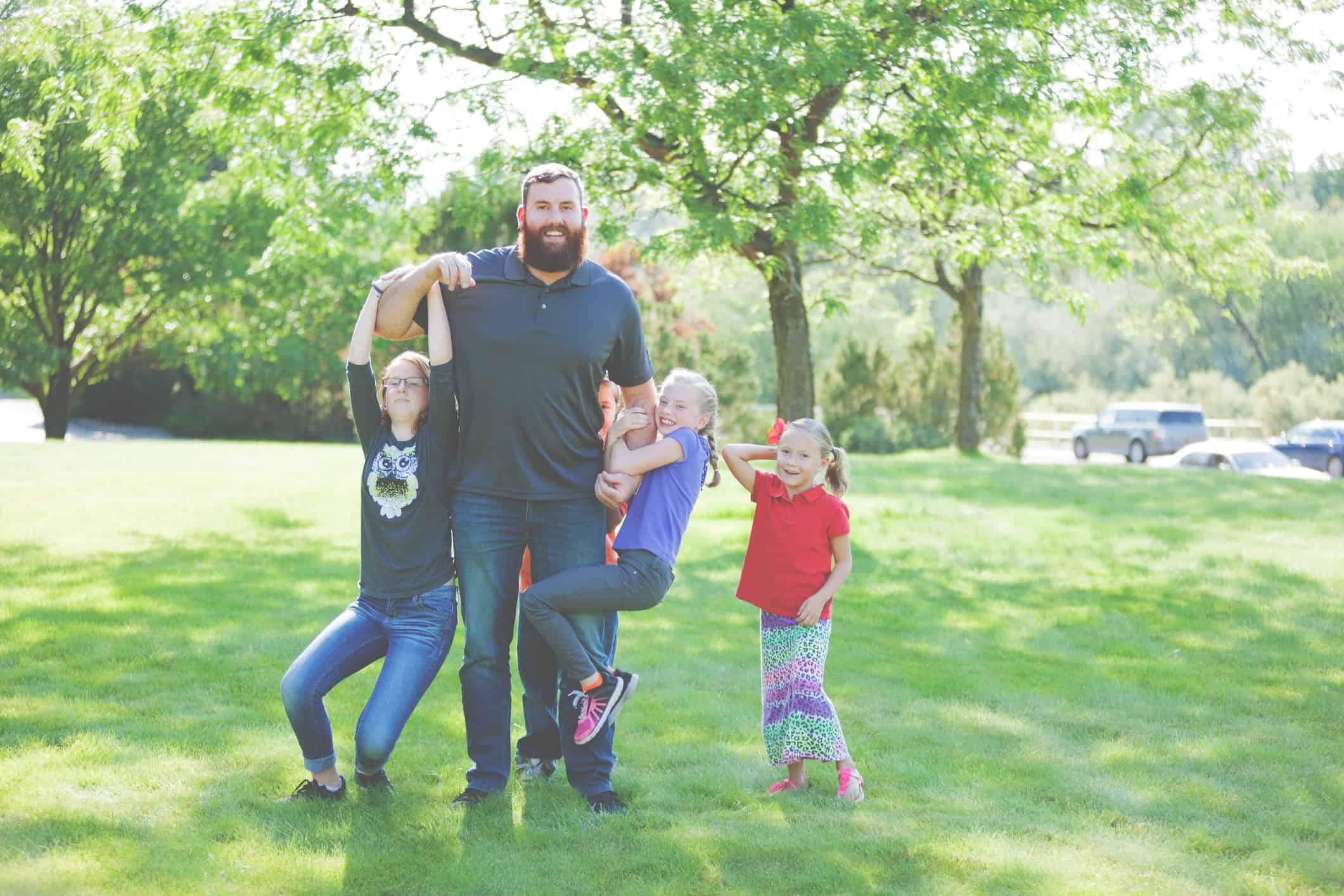 Smiling dad with his son and three daughters in a park; one daughter is hanging off each of his arms