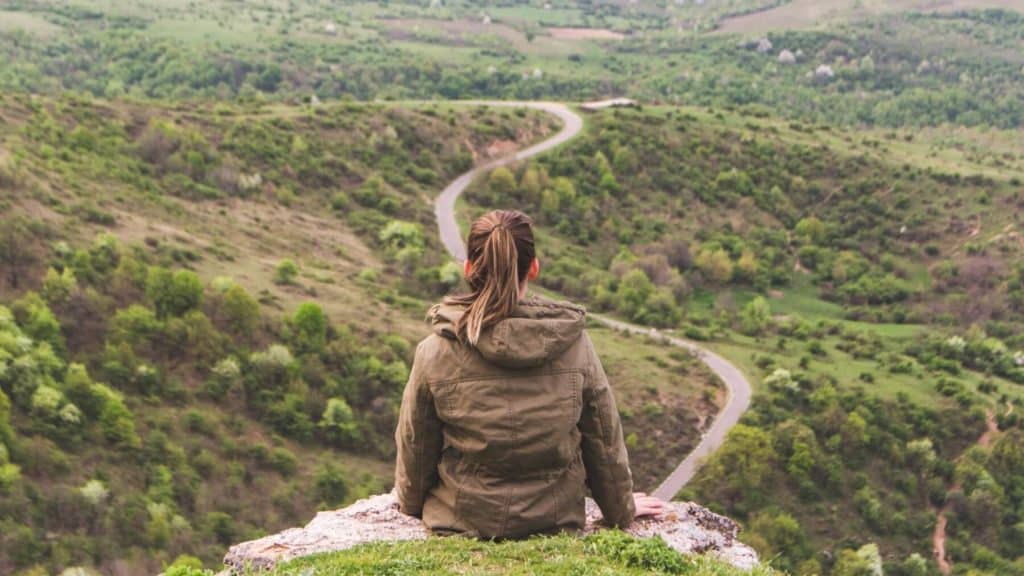 Shown from behind, a young woman sitting on a high hill overlooking a green valley with a winding road