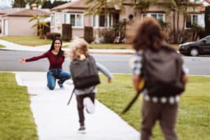 Two school kids meeting their cheerful mom outside. One is running toward her as she kneels down with outstretched arms