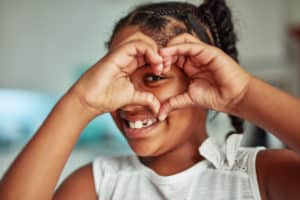 Close up of smiling black girl with one of her eyes looking through her hands which are in a heart shape