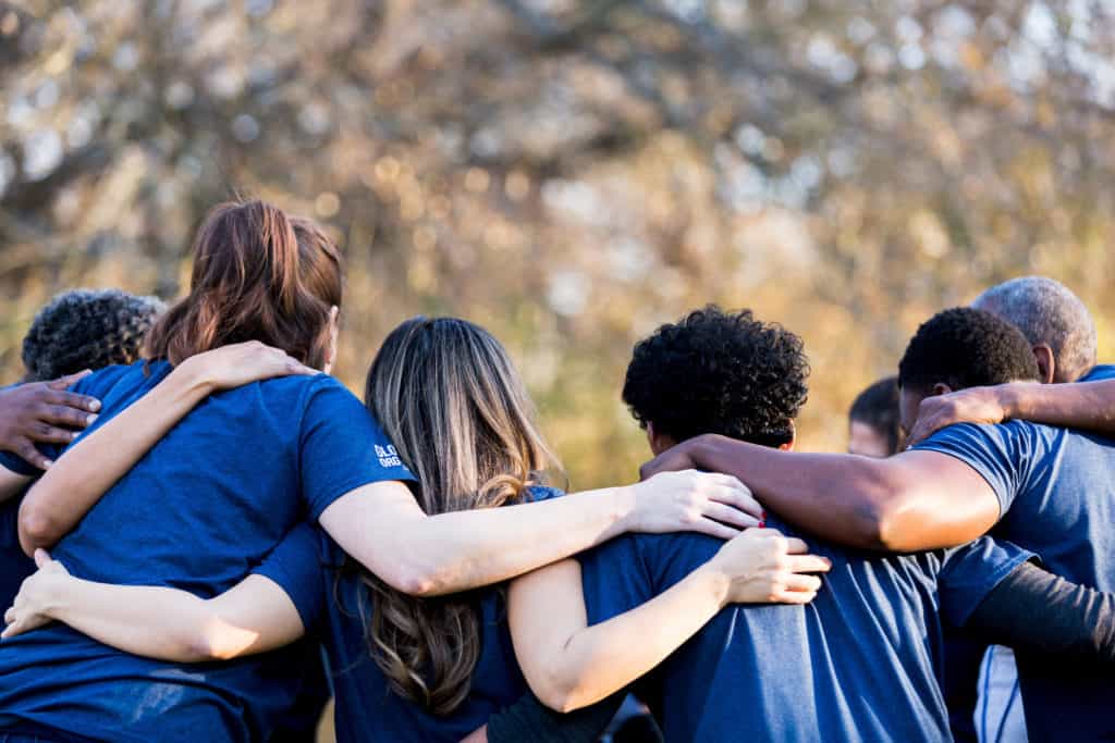 Shown from behind, a team of people in a huddle outside