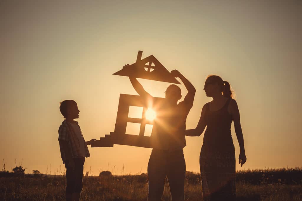 Silhouette of mom and young son holding cardboard cutout of incomplete house while dad attaches the roof