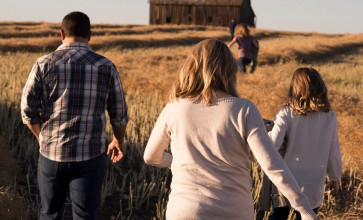 Shown from behind, a family walking through a wheat field toward a barn in the distance