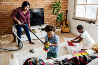 Black family cleaning their living room. Mom is vacuuming while her two young kids are putting toys away.