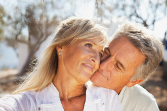 Older middle age couple standing outside. He's hugging her from behind and leaning in to her cheek for a kiss