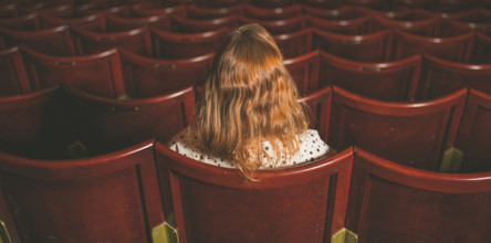 Shown from behind, a lone woman sitting in an empty theater