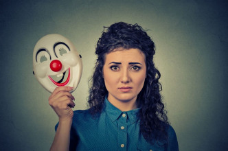Serious-looking woman holding a smiling clown mask near her face